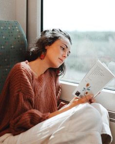 a woman sitting on a train reading a book and looking out the window with her eyes closed