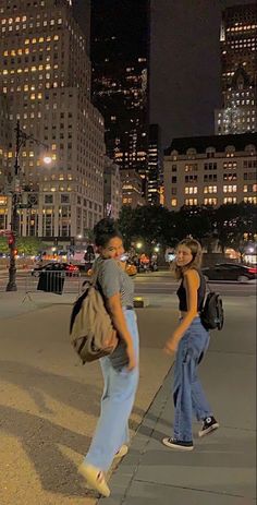 two women walking down the sidewalk in front of some tall buildings at night with lights on