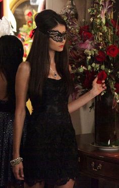 a woman in a black dress and mask holding a red rose with her right hand