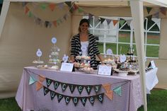 a woman standing behind a table with cupcakes and cake on it in front of a tent