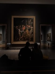 two people are sitting on a bench looking at paintings in an art gallery, with one person taking a photo