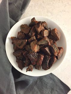 a white bowl filled with beef pieces on top of a gray cloth next to a knife