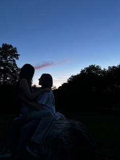 two people sitting on top of a rock under a blue sky at night with trees in the background