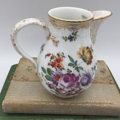 a white and gold decorated pitcher sitting on top of a book with flowers painted on it