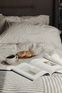 an open book, cup of coffee and some bread on a bed with white sheets