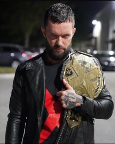 a man in black leather jacket holding up a gold and silver wrestling belt on street