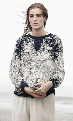 a woman standing on the beach with her hair blowing in the wind and holding a bag