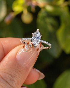 a hand holding a diamond ring in front of a bush with green leaves behind it