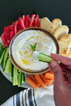 a person dipping dip into a bowl with vegetables and crackers on the side,