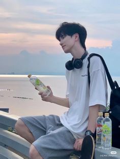 a young man sitting on a bench next to the ocean with headphones and eating food