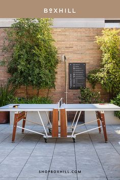 A white folding outdoor ping pong table set up in a courtyard with brick wall surrounded by greenery Folding Ping Pong Table, Pingpong Table, Iroko Wood, Ping Pong Tables, Metal Workshop, Foosball Table, Pong Table, Outdoor Games