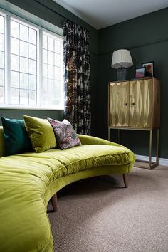 a green couch sitting in front of a window next to a dresser and lamp on top of a carpeted floor