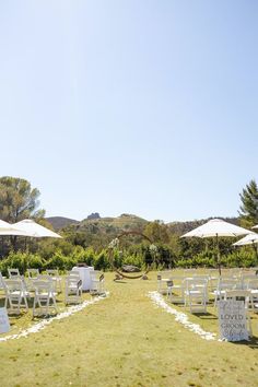 an outdoor wedding setup with white chairs and umbrellas in the grass, surrounded by greenery