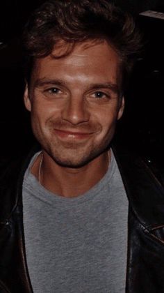 a man in a leather jacket smiling at the camera while wearing a gray shirt and grey t - shirt