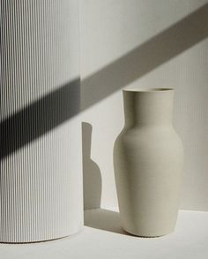 a white vase sitting in front of a wall next to a tall cylindrical object on the ground