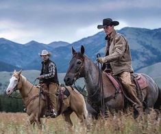 two men riding horses in a field with mountains in the background