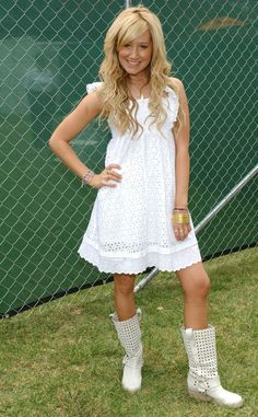 a woman standing in front of a fence with her hands on her hips and wearing white boots