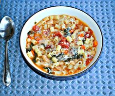 a white bowl filled with pasta and vegetables next to a spoon on a blue place mat