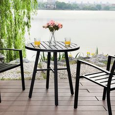 two chairs and a table on a wooden deck overlooking the water with flowers in vases