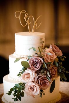 a white wedding cake with pink roses and greenery on top is decorated with the initials monogram