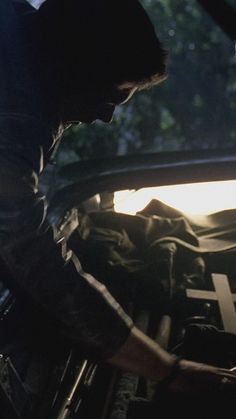 a man is working on the hood of a car with sunlight shining through trees in the background