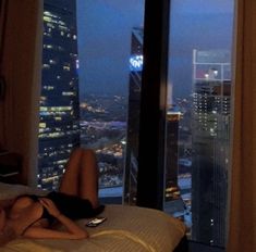 a woman laying on top of a bed in front of a window with city lights