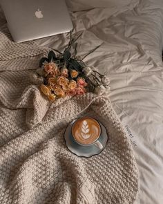 a cup of coffee sits on a bed next to an apple laptop and some flowers