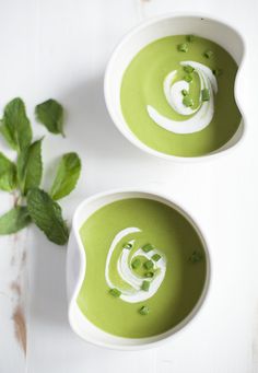 two bowls filled with green soup on top of a white table next to some leaves