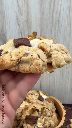 a hand holding a chocolate chip cookie with marshmallows on it and a bowl of cookies in the background