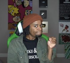 a man with dreadlocks sitting in an office wearing a black shirt and green jacket