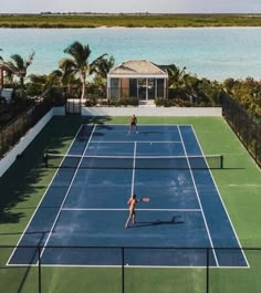 two people playing tennis on a blue and green court near the water with palm trees