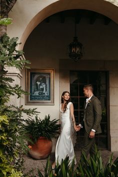 a bride and groom holding hands in front of a building with plants on either side