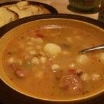 a bowl of soup with meatballs, potatoes and bread on a table next to a bottle of beer