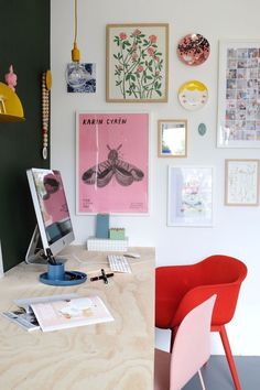 a desk with a computer and various pictures on the wall above it, along with a red chair