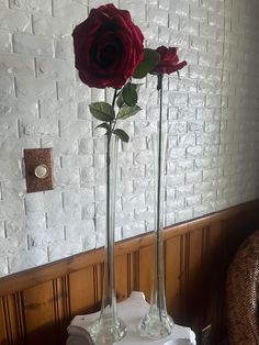 two tall vases with red roses in them on a table next to a brick wall