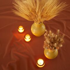 three yellow vases filled with candles on a red cloth covered tablecloth next to some wheat stalks