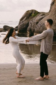 a man and woman are holding hands on the beach near the water with rocks in the background