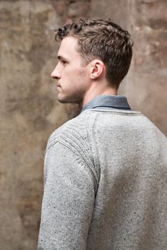 a man standing in front of a stone wall wearing a gray sweater and black pants