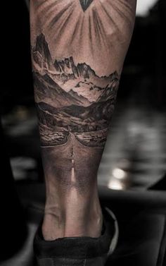 a man's leg with mountains and a cross tattoo on his left calf area