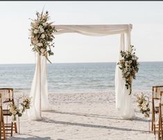 an outdoor wedding setup on the beach with white draping and floral arrangements, along with gold chairs