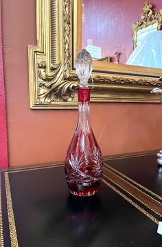 a red glass bottle sitting on top of a black table next to a gold framed mirror