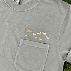 a gray t - shirt with daisies embroidered on the front and back pocket, laying in grass