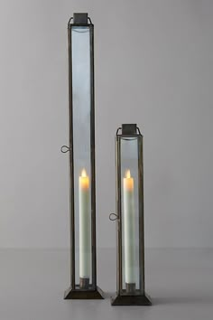 two tall metal candles stand next to each other on a gray surface with one lit candle in the middle