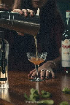 a woman pouring a drink into a glass on top of a wooden table next to bottles