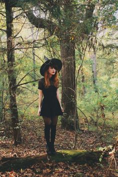 a woman standing in the woods wearing a black dress and hat with her hands on her hips