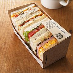 a sandwich in a cardboard box on a wooden table next to a cup and saucer