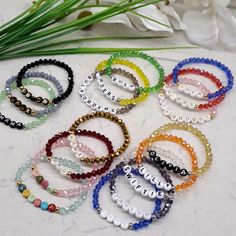 six bracelets with different colored beads on a marble surface next to flowers and greenery