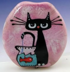 a black cat holding a tea pot on top of a pink rock with a blue fish in it's mouth