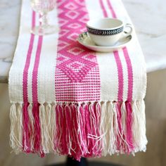 a pink and white table runner with tassels on it, next to a coffee cup