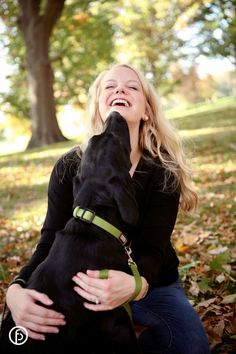 a woman sitting on the ground with her dog in front of some trees and leaves